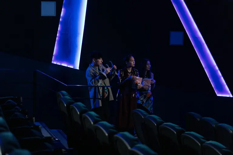 Three friends standing in a dimly lit cinema holding popcorn, enjoying a movie night.