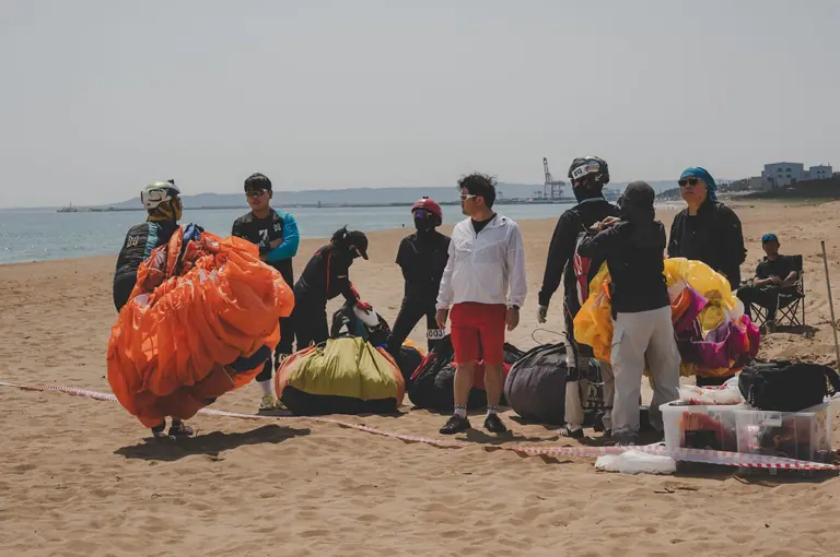 Group of adult paragliders preparing on a sunny beach, with parachutes and gear.