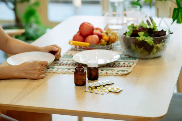 From above of crop anonymous person sitting at table with pile of various medicines and bowls of fresh lettuce salad and ripe fruits in kitchen