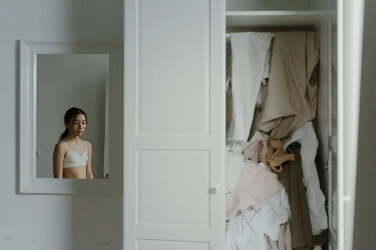 A young woman reflects in the mirror beside an unorganized closet.