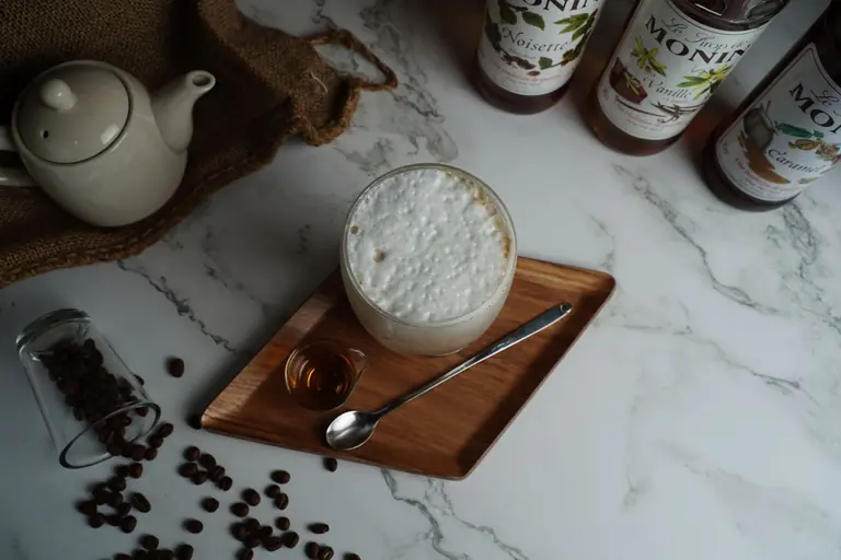 Top view of a foamy coffee drink with syrup and a spoon on a wooden tray, featuring coffee beans and syrup bottles on a marble surface.