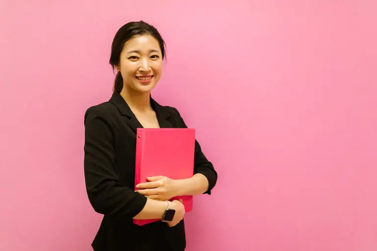 Smiling Asian woman holding a pink binder against a pink background, conveying positivity.