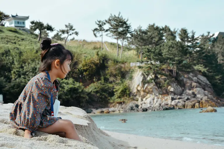 A young girl sitting on a sandy beach in South Korea, gazing at the ocean.