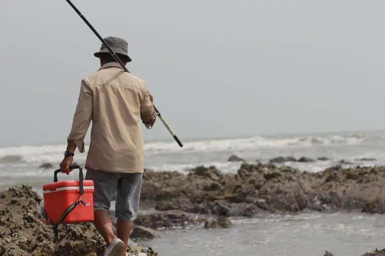 A lone fisherman walks along the rocky shore carrying fishing gear and a cooler.
