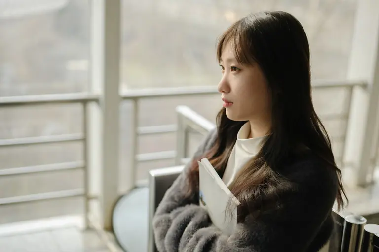 An adult woman with long black hair gazes thoughtfully out of a large window while holding a book.