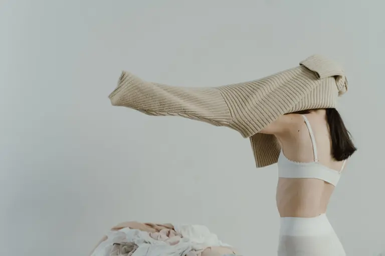 A woman wearing a white brassiere and putting on a beige sweater in a minimalist style room.