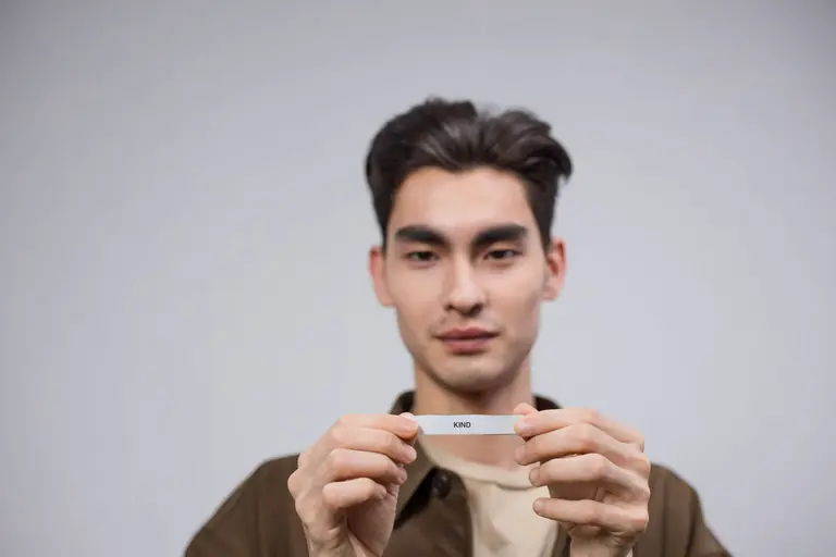 Portrait of a young man holding a strip of paper with the word 'kind' in a studio setting.