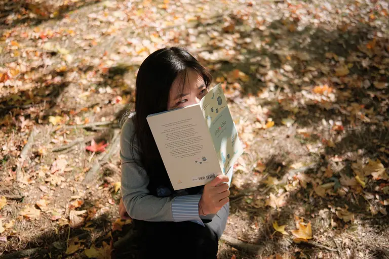 A young woman enjoys reading a book in a sunlit autumn park, surrounded by fallen leaves.