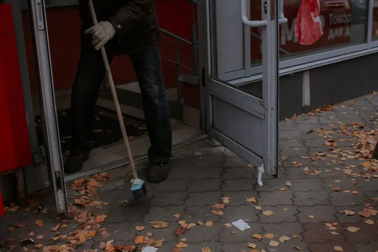 Person sweeping dry leaves outside a shop entrance during fall.
