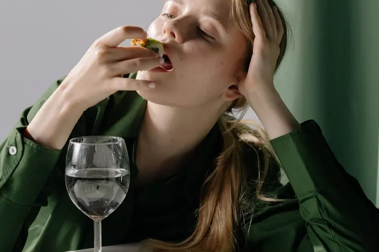 Portrait of a young woman savoring sushi with a wine glass in a relaxed, green-themed setting.