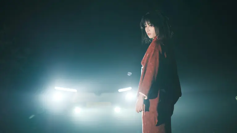 A woman in traditional clothing stands in front of car headlights at night, creating a mysterious atmosphere.