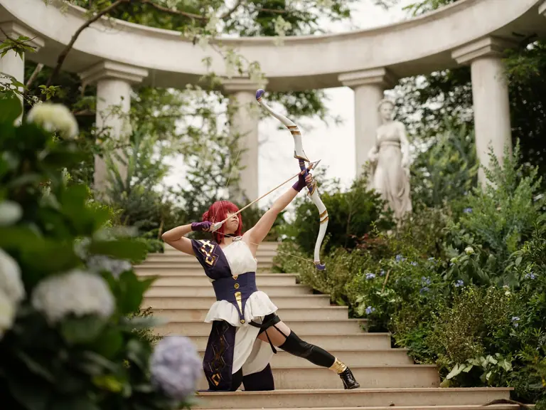 Cosplayer in archer attire performs dynamic pose in a serene garden with classical architecture.
