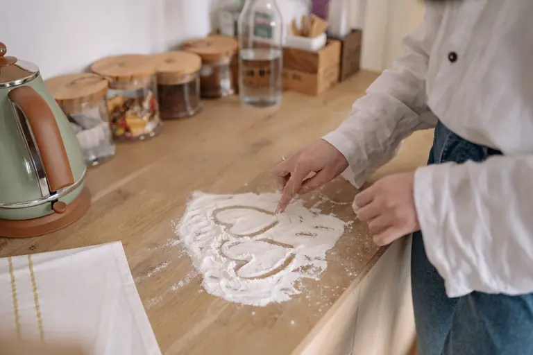 A person uses their finger to draw a heart shape in flour on a wooden kitchen counter.