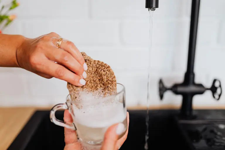 A close-up view of hands washing a glass with soap under a kitchen faucet.