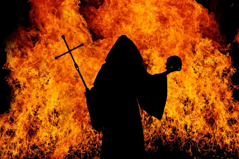 Dramatic silhouette of a figure in a ritual holding cross and skull in front of blazing flames.