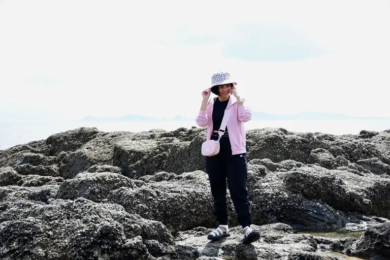 Asian woman standing on rocky shore in Vũng Tàu, Vietnam, wearing a pink jacket and summer hat.