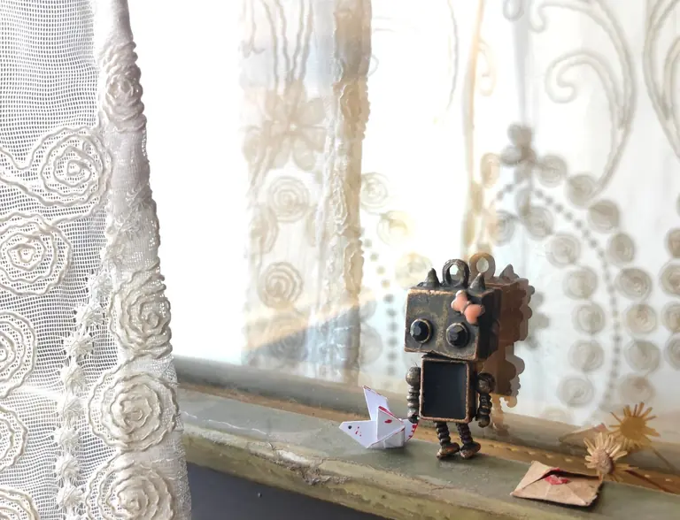A charming robot figurine with an origami heart sits on a windowsill next to lace curtains, evoking a playful scene.