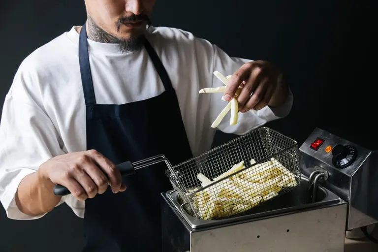 A chef handling raw french fries with a strainer basket in a deep fryer.