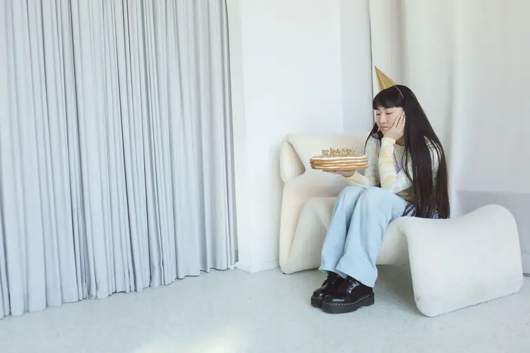 An Asian woman with long hair sits indoors, holding a birthday cake and wearing a party hat.
