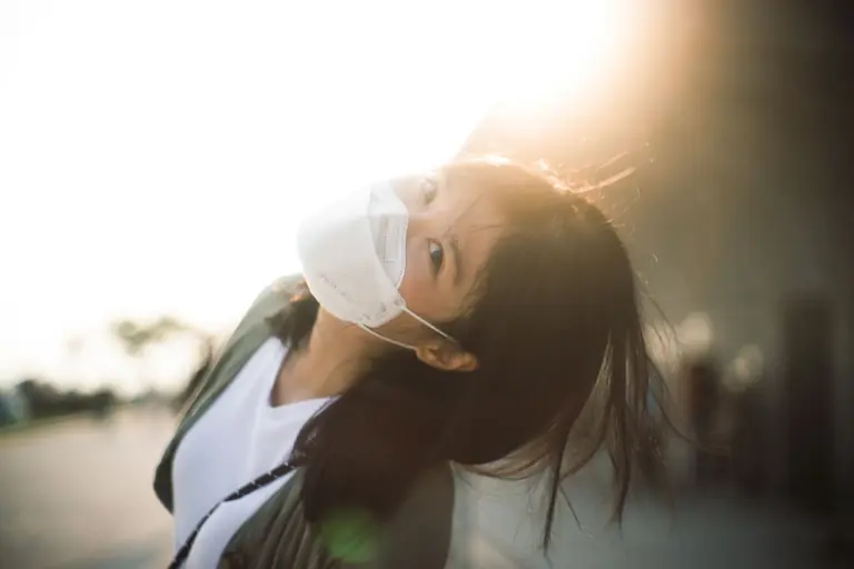 Portrait of a young woman wearing a face mask outdoors with a sunlit background.