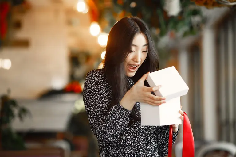 An Asian woman delightfully opens a gift box with a red ribbon indoors.