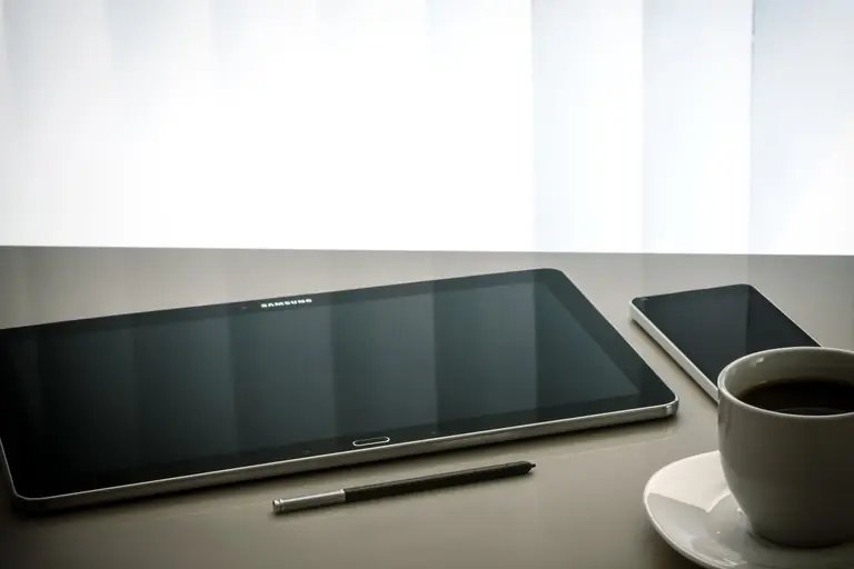 Minimalist office desk setup featuring a tablet, smartphone, pen, and coffee cup on a reflective surface.