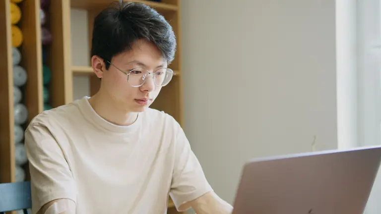 Young man focused on work, wearing glasses, using laptop in well-lit room. Ideal for productivity themes.