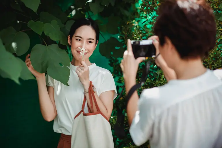 Crop anonymous lady taking photo of smiling best Asian friend with flower on camera in city park