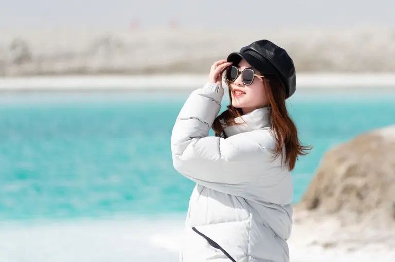 Trendy woman in a coat with sunglasses poses by a serene beach.