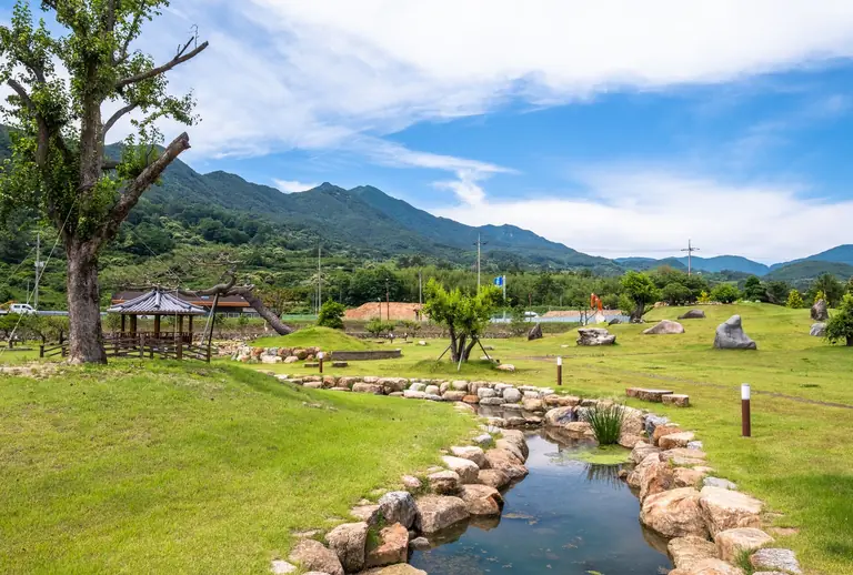 A serene park scene with a wooden gazebo, stream, and mountain view under a clear sky.