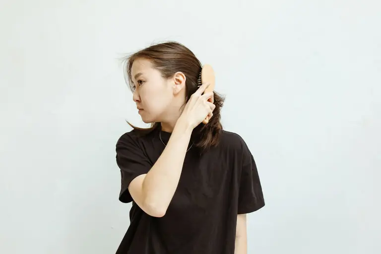 An Asian woman combs her hair in a minimalist setting with a white background.