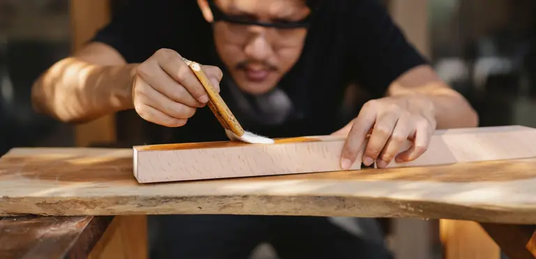 Serious crop young ethnic male carpenter in casual clothes and eyeglasses applying varnish to wooden plank with brush in workshop